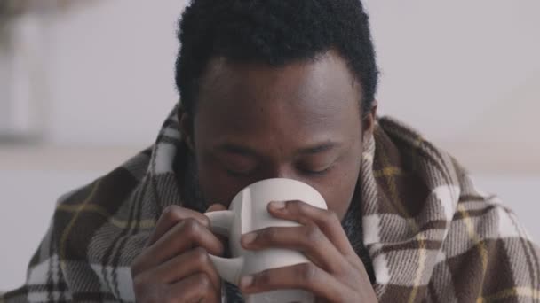 Nahaufnahme Porträt eines kranken afrikanisch-amerikanischen Typen, der in Plaid gehüllt Heiltee trinkt und traurig in die Kamera blickt - Filmmaterial, Video