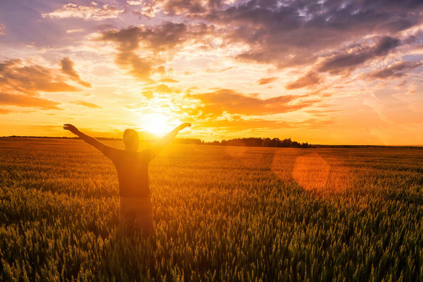Silhouette eines Mannes hebt die Hände bis Sonnenuntergang oder Sonnenaufgang auf dem Feld mit jungen Roggen oder Weizen im Sommer mit bewölktem Himmel Hintergrund.  - Foto, Bild