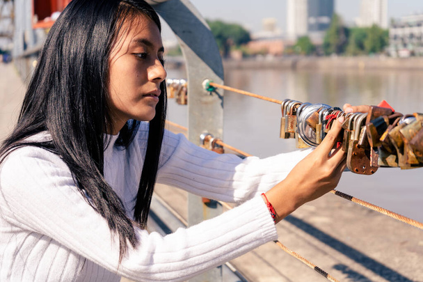 Beautiful black-haired Latin woman putting a lock on one of the bridges in a city. Concept fidelity, romantic love. - Photo, Image