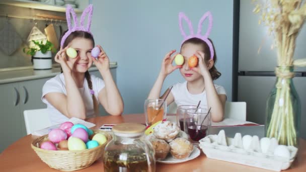Hyvää pääsiäistä. Kaksi siskoa maalaa pääsiäismunia. Onnelliset perheen lapset valmistautuvat pääsiäiseen. Söpö pieni lapsi tyttö yllään pupu korvat poseeraa värillisiä munia kameran edessä - Materiaali, video