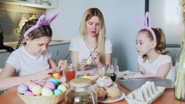 Mladá matka a její dcery malují velikonoční vajíčka. Šťastná rodina se připravuje na Velikonoce. Roztomilé holčičky nosí králičí uši o Velikonočním dni. - Záběry, video