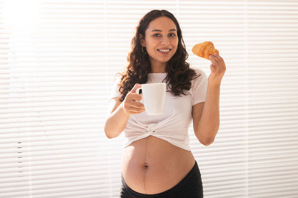 Hermosa mujer embarazada sosteniendo croissant y taza de café en sus manos durante el desayuno de la mañana. Concepto de buena salud y actitud positiva mientras espera al bebé - Foto, Imagen