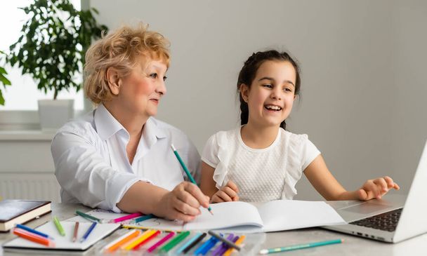 Γυναίκα δάσκαλος ή ανάδοχη μητέρα βοηθώντας χαριτωμένο καυκάσιο κορίτσι σχολείο κάνει την εργασία κάθεται στο τραπέζι. Διαφορετική νταντά και το παιδί μαθαίνουν γραφή στο σημειωματάριο μελέτη στο σπίτι. - Φωτογραφία, εικόνα