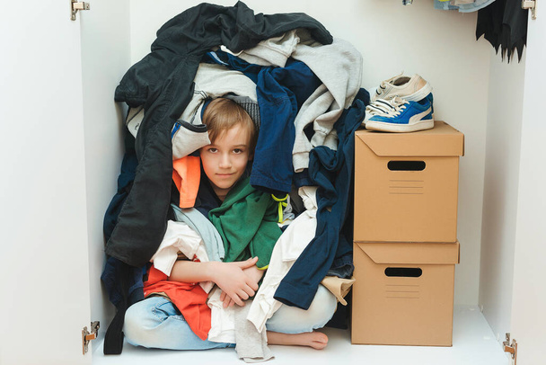 Kid hiding among messy clothes inside closet. Organization and storage of clothes at home. Mess in the wardrobe. Boy with messy colorful clothing at room. Untidy clutter clothing closet. - Photo, Image