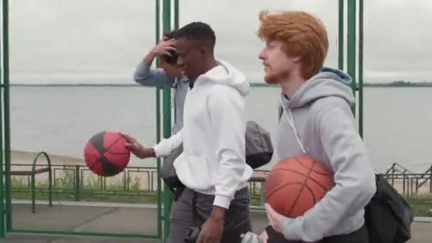 Tracking shot van drie jonge mannen in hoodies dragen sporttassen, waterflessen en basketballen en wandelen op outdoor basketbalveld op sombere dag - Video