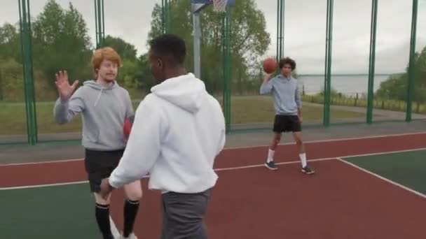 Ruční sledování záběr veselý mladý muž dává high-five na svého přítele a skákání basketbal na venkovním hřišti v ponurý den - Záběry, video