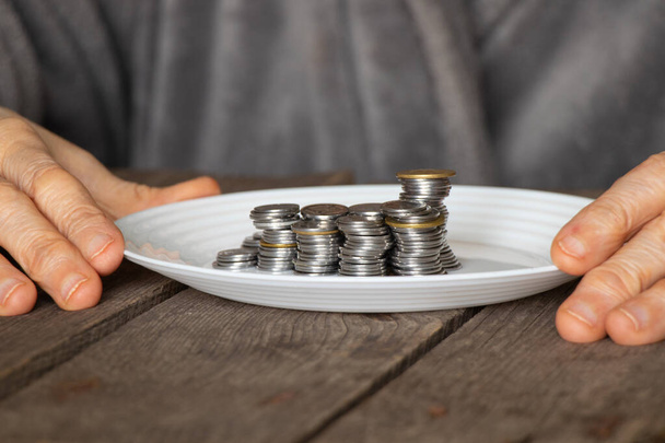 una pila de monedas en un plato blanco sobre una vieja mesa de madera y viejas manos femeninas vertían un plato en casa en la cocina - Foto, Imagen