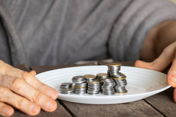 una pila di monete su un piatto bianco su un vecchio tavolo di legno e vecchie mani femminili versato un piatto a casa in cucina - Foto, immagini
