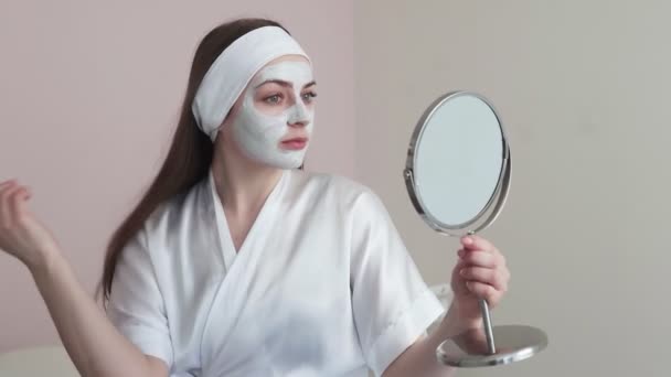 Νεαρή γυναίκα σε ινστιτούτο αισθητικής ομορφιάς με πήλινη μάσκα στο πρόσωπο κοιτάζει τον καθρέφτη - Πλάνα, βίντεο