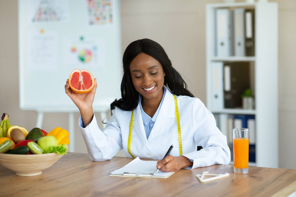 Ελκυστική Αφροαμερικανός διατροφικός σύμβουλος κρατώντας γκρέιπφρουτ, κάνοντας σχέδιο γεύματος για τον πελάτη σε κλινική απώλειας βάρους - Φωτογραφία, εικόνα