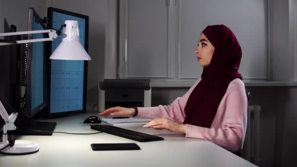 Una donna in hijab si siede in ufficio davanti al computer e digita sulla tastiera - Filmati, video