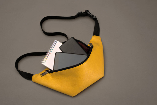 アクセサリーと濃いグレーの背景に女性と男性のための黄色のベルトバッグ:ノートパッド、パワーバンクと電話。コピースペース。最上階だ。トレンドカラー2021年パントン. - 写真・画像