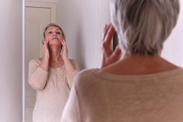 Vanhukset valkoihoiset naiset, joilla on lyhyet harmaat hiukset rennoissa vaatteissa, huolehtivat kasvojen ihosta, tekevät itsestään hieronnan.. - Valokuva, kuva