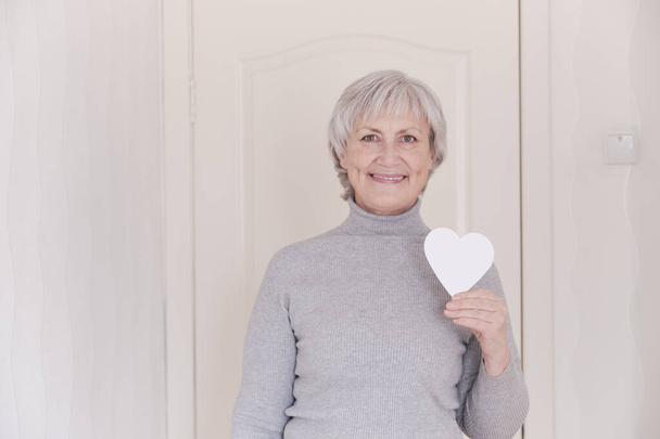 Egy mosolygó idős, fehér nő portréja, rövid ősz hajjal a kezében, kezében egy kis fehér szívvel.. - Fotó, kép
