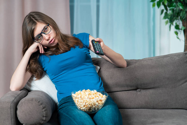 Bored girl in glasses, young tired woman with boring face is sitting on couch or sofa at home in living room with TV remote in hand, watching TV, boring movie, uninteresting film with plate of popcorn - Фото, изображение