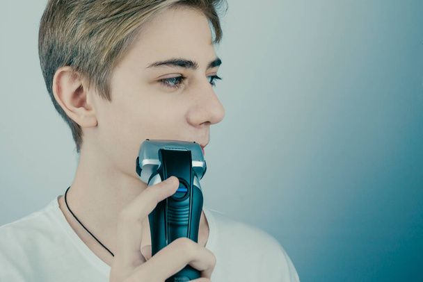 Portrait of a handsome smiling teenager in a white T-shirt shaves for the first time, shaves off his mustache and beard with an electric razor - Photo, Image