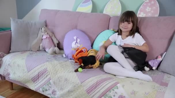 Urocza dziewczynka przytula swojego zwierzaka w domu na kanapie. Dzieciak przygotowuje się do Wielkanocy w domu podczas kwarantanny. Słodki pies - Materiał filmowy, wideo