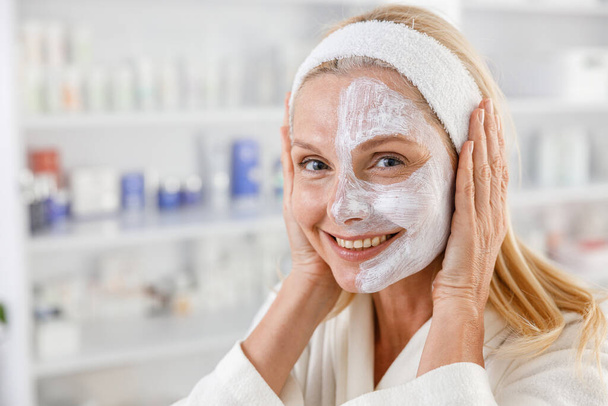 Femme âgée avec masque cosmétique sur demi-visage au salon de beauté - Photo, image