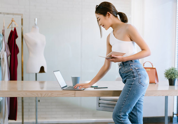 Portrait of young Asian woman fashion designer using on laptop computer and holding notebook check online orders to prepare sell to customers. Startup Small business owner, SME e-commerce concept - Photo, Image