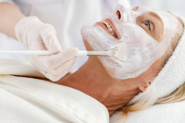 Nahaufnahme Foto einer Kosmetikerin, die Tongesichtsmaske auf das Gesicht einer Seniorin aufträgt - Foto, Bild