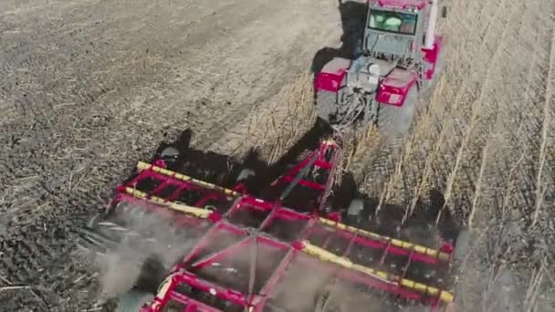 Luftaufnahme eines roten Traktors mit Eggenanlage, der Boden auf einem bewirtschafteten Feld pflügt, dahinter eine Säule von Staubspuren, die Erde für die Anpflanzung neuer Pflanzen vorbereitet - Filmmaterial, Video