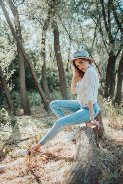 A vertical shot of a young stylish female sitting on a tree stump in a forest - Фото, изображение