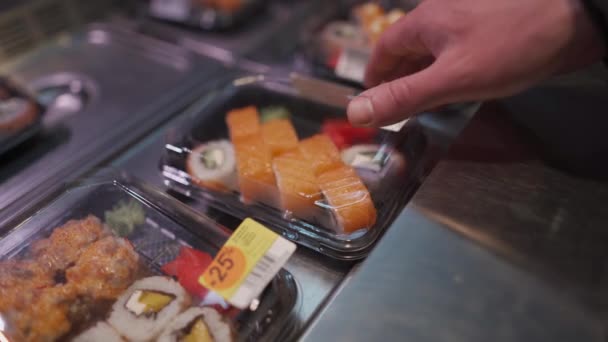 Λευκός άντρας με προστατευτική μάσκα στο πρόσωπό του διαλέγει σούσι σε κουτί για να πάει στο σούπερ μάρκετ κατά τη διάρκεια της καραντίνας και της επιδημίας του κορωναϊού. Ιαπωνικό φαγητό για πακέτο. Υγιεινά τρόφιμα, κουτί φαγητού - Πλάνα, βίντεο