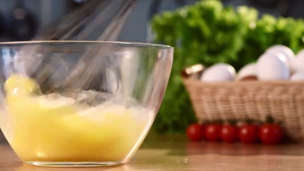 Close-up shot of whisking fresh raw chicken eggs in a glass bowl with pouring milk in process. Shiny colorful background with white eggs and green salad. - Materiał filmowy, wideo