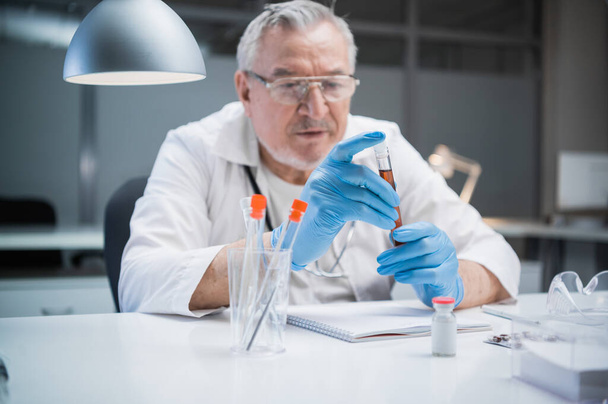 Un asistente de laboratorio médico examina las propiedades de la vacuna producida. Él sostiene una botella con una muestra de sangre que contiene una vacuna contra el virus covid-19 - Foto, imagen