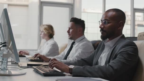 Снимок средней замедленной съемки трех мультиэтнических разработчиков программного обеспечения в формальной одежде, проводящих рабочий день в офисе, работающих на настольных компьютерах - Кадры, видео