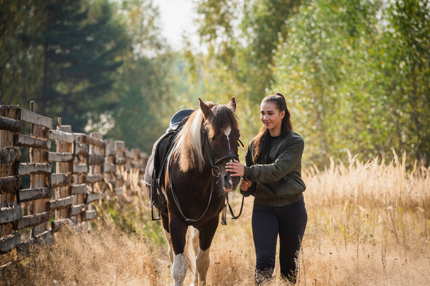 Молодая девушка ведет свою лошадь за уздечку по тропинке вдоль забора - Фото, изображение