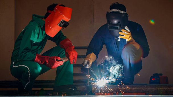 Zwei Ingenieure Mechaniker sitzen und arbeiten in der Werkstatt einer Fabrik. Sie helfen sich gegenseitig, nachts mit einem Schweißgerät ein Stück Metalldraht zu schweißen. - Foto, Bild