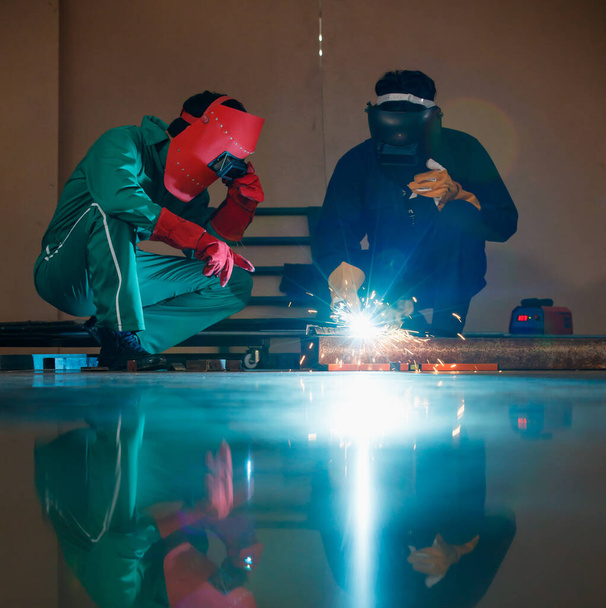 Dos ingenieros mecánicos sentados y trabajando en un taller de una fábrica. Se están ayudando mutuamente a soldar una pieza de varilla de metal con una máquina de soldadura por la noche. - Foto, Imagen