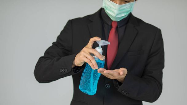 スーツを着た実業家がアルコールゲルを保持する保護衛生マスクを着用し、コロナウイルス保護のための手を洗浄し、滅菌する方法を実証するために手に注ぐ. - 写真・画像