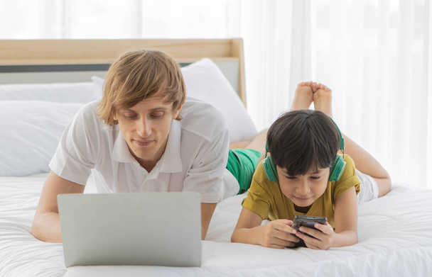 白人の父と混合レースの息子はベッドの上に横たわって、デバイスとインターネットに接続します。父はノートパソコンを使い子供はスマートフォンを使います. - 写真・画像