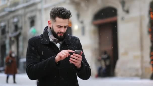 Muž stojí na centrálním náměstí ve sněhu. Píše sms na smartphone a usmívá se. 4K - Záběry, video