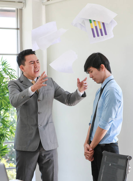 Aasialainen liikemies puvussa heittää paperitöitä ilmaan ja syyttää nuorta työntekijää vihasta ja vakavasta eleestä. Toimistotyön ongelma ja työn laadun konflikti käsite - Valokuva, kuva
