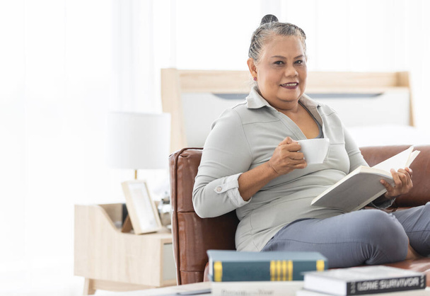 Starší starší šedé vlasy asijské žena drží šálek kávy a čtení knihy na pohovce s klidem a soustředění póza. - Fotografie, Obrázek