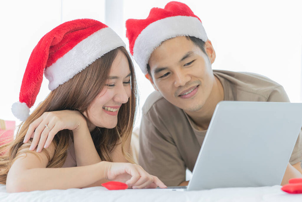 jong aziatisch paar dragen rood en wit santa hoed kijken naar elkaar glimlachend en liggend op schoon wit laken bed omgeven door mini rood hart, laptop en rode geschenkdoos. - Foto, afbeelding