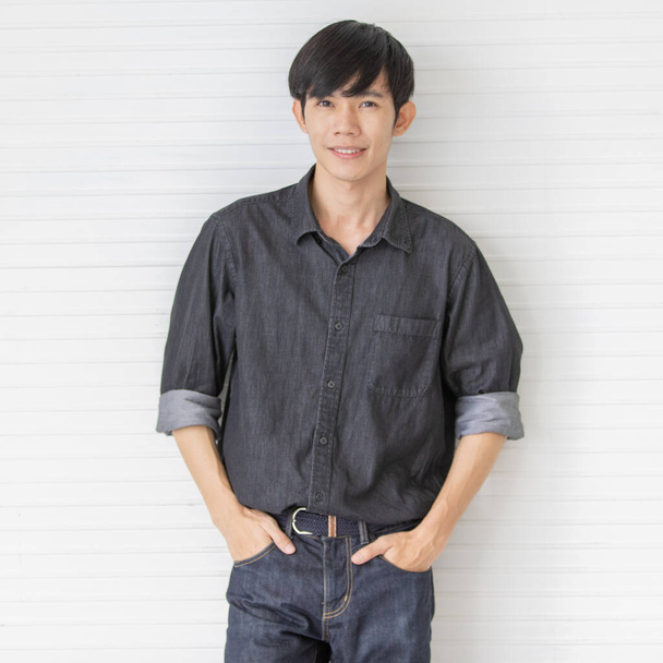 Zdjęcie portretowe azjatyckiego modelu męskiego z krótkimi czarnymi włosami w czarnej koszuli stojak uśmiecha się przed białym paskiem tła umieścić ręce w kieszeni dżinsów. - Zdjęcie, obraz