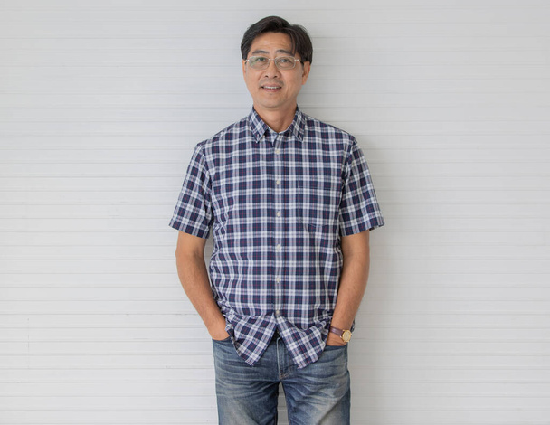 Zdjęcie portretowe azjatyckiego mężczyzny w średnim wieku z krótkimi czarnymi włosami w niebieskiej koszuli w kratę z uśmiechniętym stojakiem przed białym paskiem tła włożył ręce do kieszeni dżinsów. - Zdjęcie, obraz