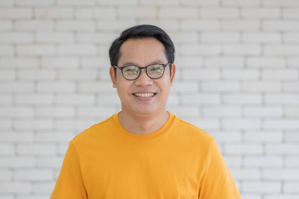 Retrato de hombre asiático adulto con camiseta amarilla y anteojos de pie y sonriendo mirando a la cámara con amigable y seguro de sí mismo. - Foto, imagen