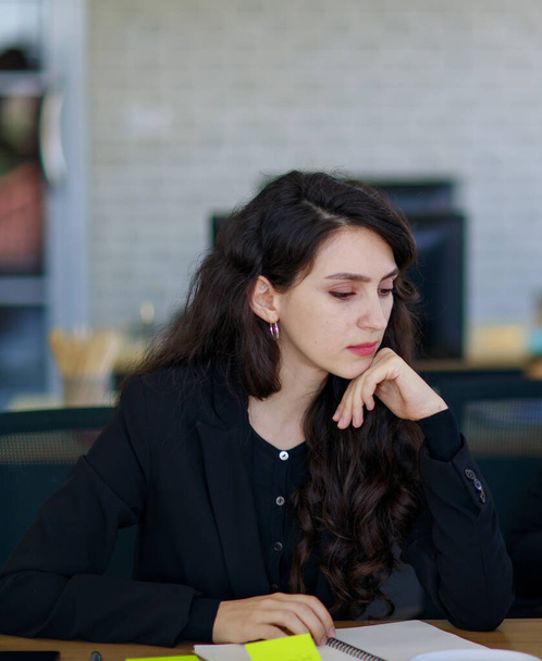 Attraktive ernsthafte kaukasische Geschäftsfrau im schwarzen Anzug mit stylischer Frisur, die nach unten schaut, Tisch mit Kaffeetasse, Farbmarker und Notizblock konzentriert und darüber nachdenkt, wie man das Problem löst - Foto, Bild