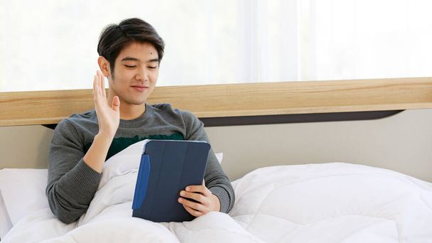 Asyalı genç adam yatak odasında oturuyor ve tablet bilgisayar ekranına el sallıyor birini selamlıyormuş gibi görünüyor.. - Fotoğraf, Görsel