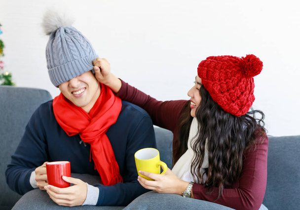 Retrato de linda joven sonriente pareja amante asiática en sudadera, bufanda, sombrero de punto relajante en el sofá con fondo de árbol de Navidad. La esposa trata de cubrir los ojos de su marido sombrero. - Foto, imagen