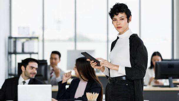 Χαριτωμένη και όμορφη επιχειρηματίας με κοστούμι φόρεμα στέκεται στο γραφείο και χρησιμοποιώντας έναν υπολογιστή tablet με αυτοπεποίθηση και κομψή στάση. - Φωτογραφία, εικόνα