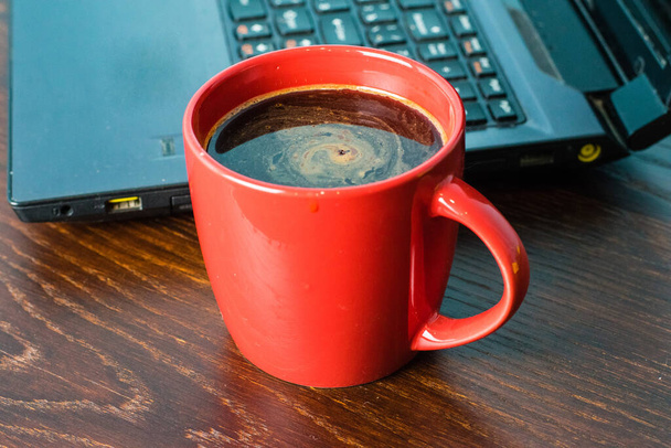 Kopje koffie en laptop op de achtergrond op een bruine houten tafel. Begrip werk op afstand of opleiding, het begin van de werkdag op kantoor - Foto, afbeelding