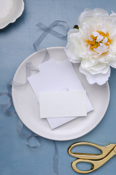Верхний вид на тарелку и белый конверт, пустая бумажная карточка на нем, белый пион, золотые ножницы и лента на синем столе - Фото, изображение