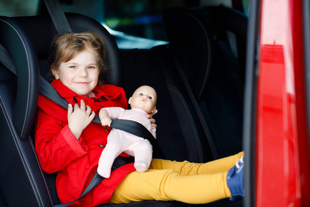 Toddler girl sitting in car seat, holding favourite doll toy and looking out of the window on nature and traffic. Little kid traveling by car. Child safety on the road. Family trip and vacations - Photo, Image