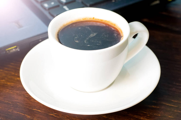Kaffee in einer weißen Tasse und ein Laptop im Hintergrund auf einem braunen Holztisch. Konzept der Fernarbeit oder Schulung, der Beginn des Arbeitstages im Büro - Foto, Bild